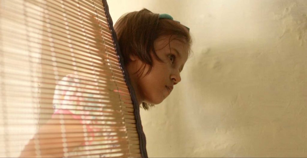 A little girl peeks around a bamboo blind in a room with warm colors