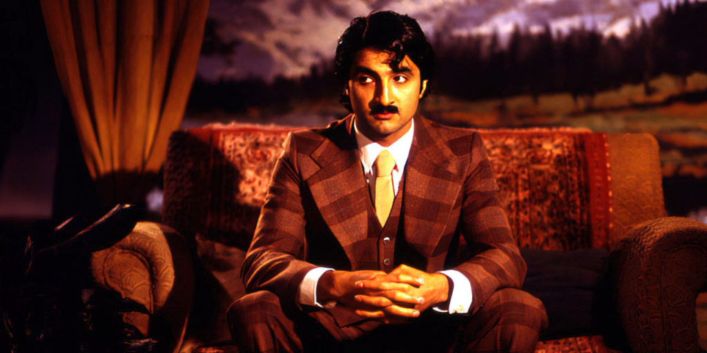 Romantic moustachioed man wearing a plaid suit sits in a moody, dark-lit setting