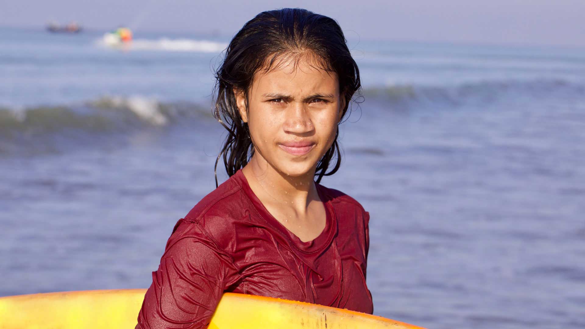 A young woman with wet hair, holding her surfboard, looks into the the camera with a confident and determined expression.
