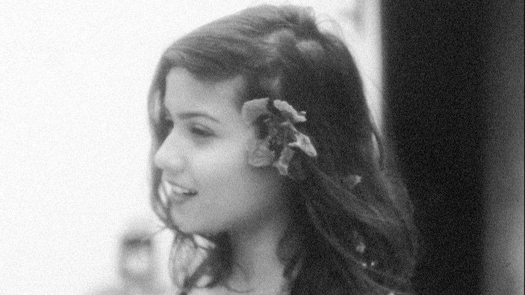 Image from film A Night of Knowing Nothing. A grainy black and white image of a young South Asian woman looking happy with a flower in her hair.