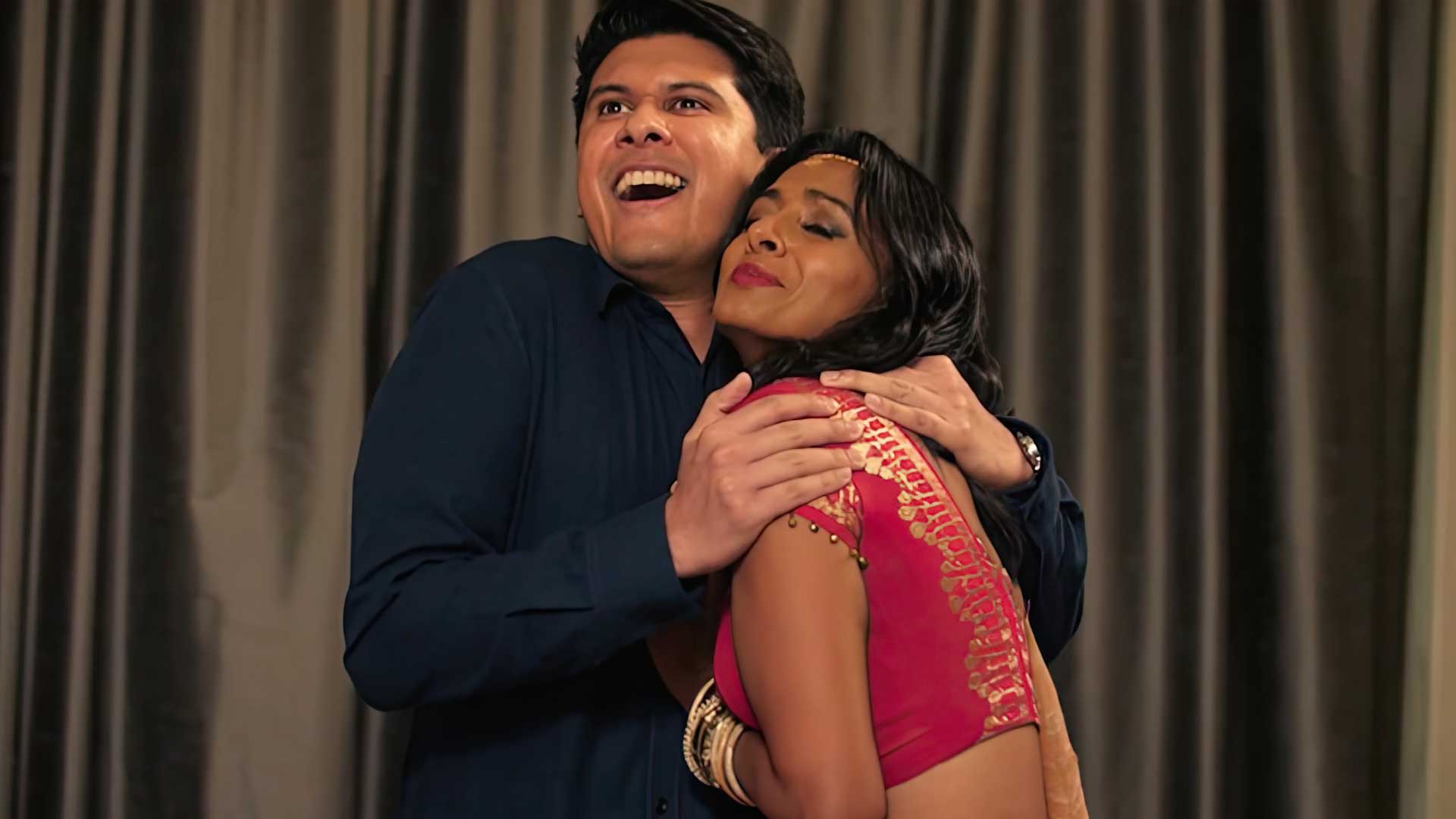 A man and woman dressed like Golden Age Bollywood actors embrace comically in front of a simple taupe-colored curtain.