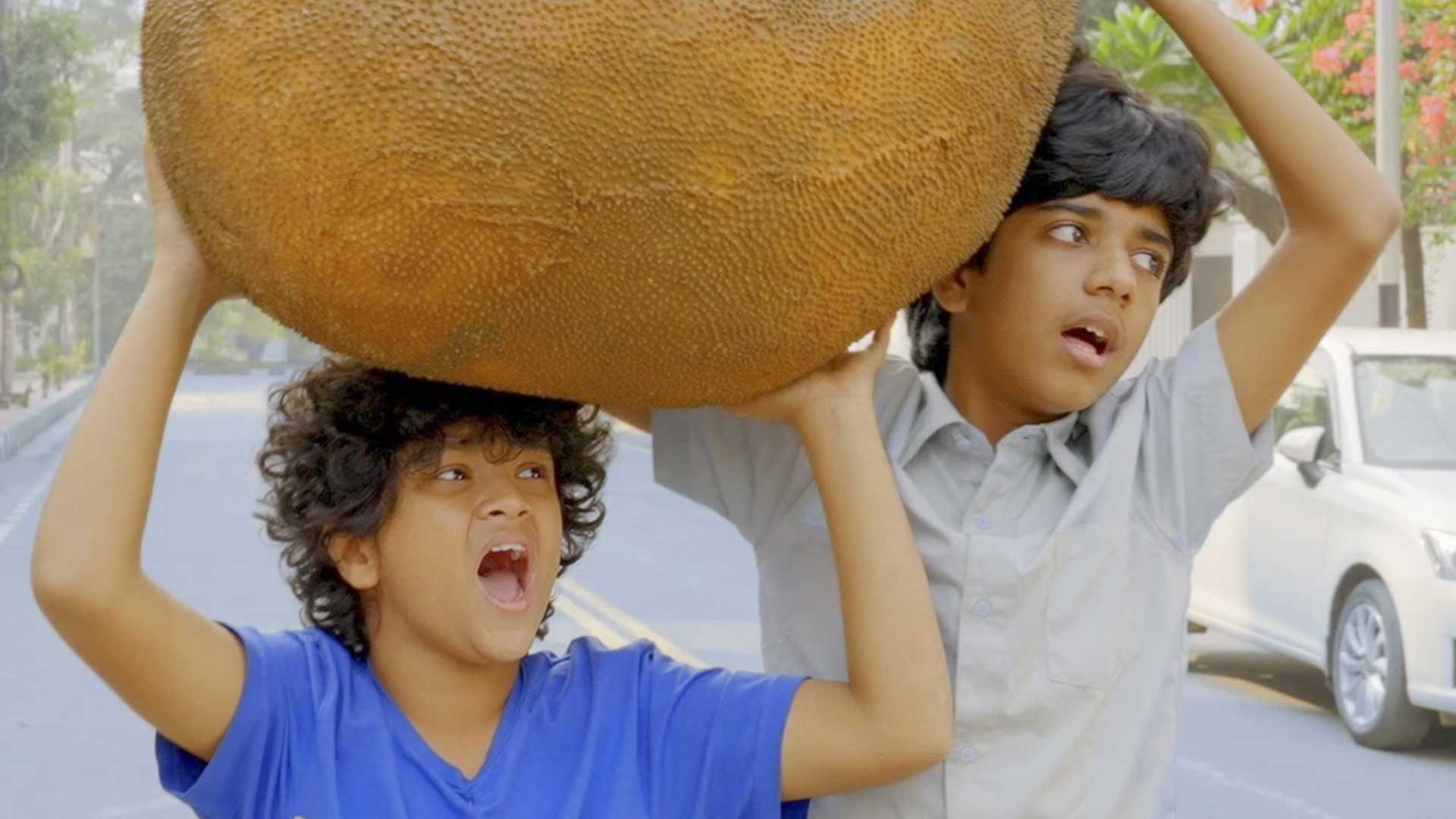 Two young boys in Bangladesh are surprised by something outside of the frame as they carry an enormous jackfruit.