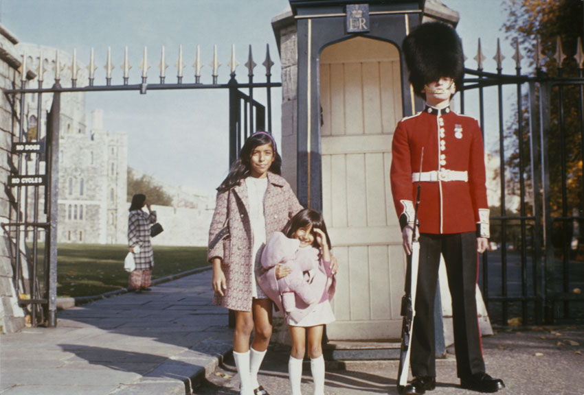 A late-1960s photo showing two young girls of Indian heritage, wearing respectable outfits with knee socks, posing next to a Beefeater in a red uniform tunic and giant bearskin hat outside Buckingham Palace in London.