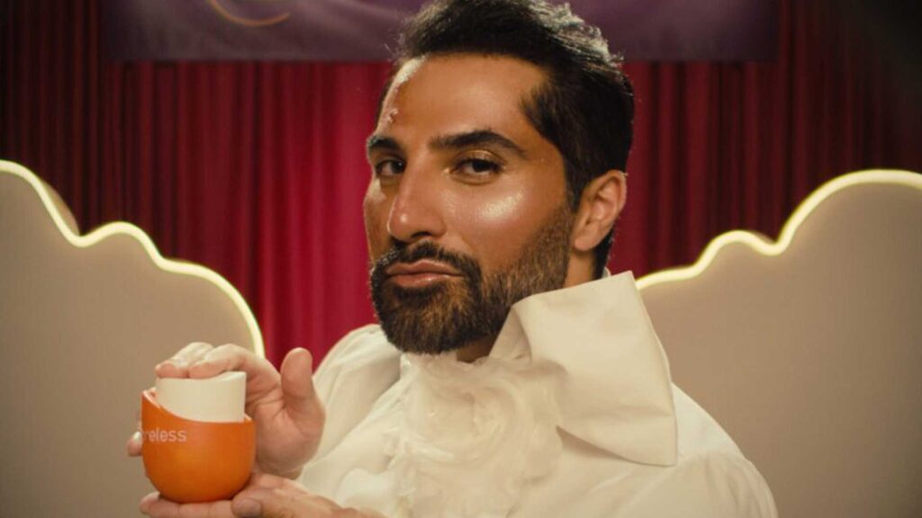 A South Asian man with a well-groomed beard and eyebrows, wearing a ruffled, cream shirt and standing in front of a displaly of stylized fake clouds, holds up a modern, streamlined container. Still from the film Poreless.