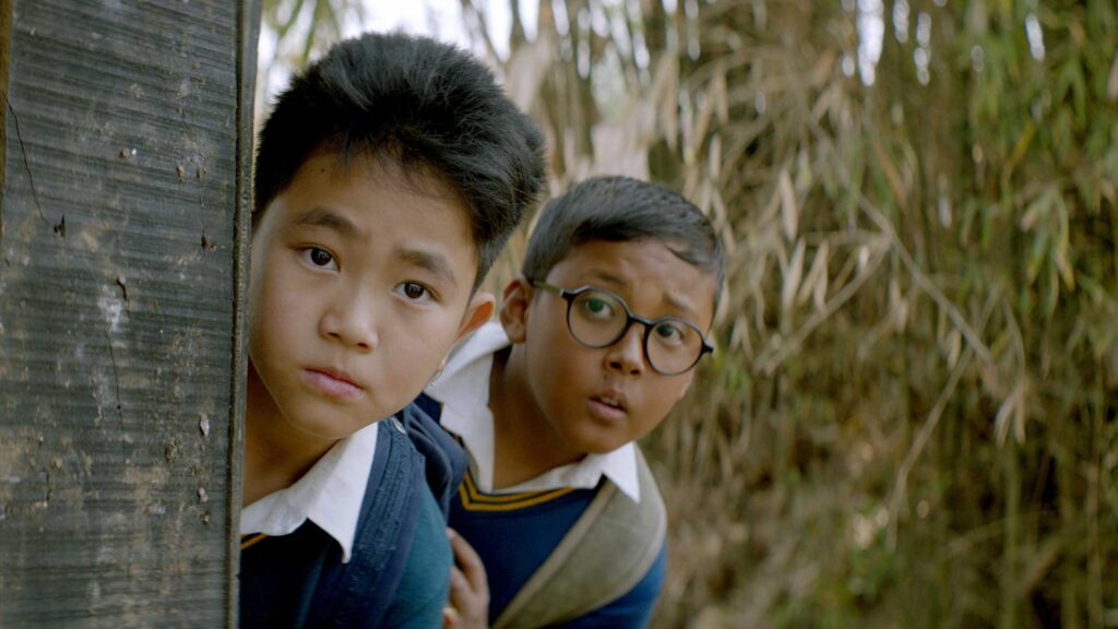 two young boys in Manipur, India, out on a secret adventure, peer around a wooden wall.