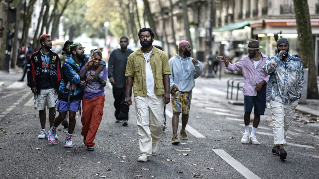 A group of young people of Sri Lankan descent stroll toward the camera in a neighborhood of Paris. The central figure is a young man who wears an intense, troubled expression, while his friends are laughing obliviously.