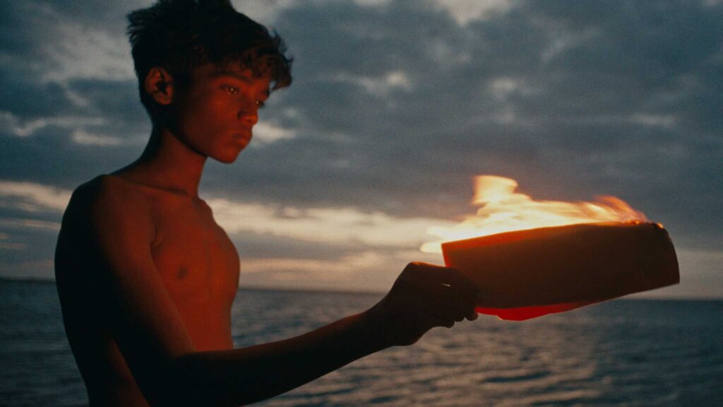 An adolescent boy in Sri Lanka stands in front of the sea at sunset, with a moody cloud-filled sky in the background. His face and bare chest are illuminated by a warm glow from the letter he is holding, which is aflame.