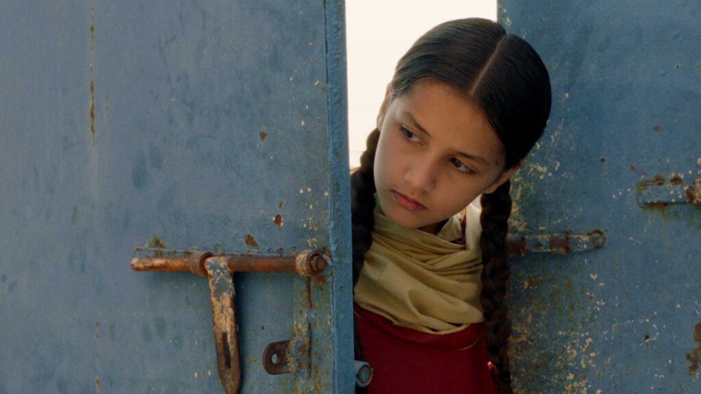 Stiil from film Ghost School, SFISAFF 2026, shows a girl in braids peeking through a rustic pale blue door, in dim lighting