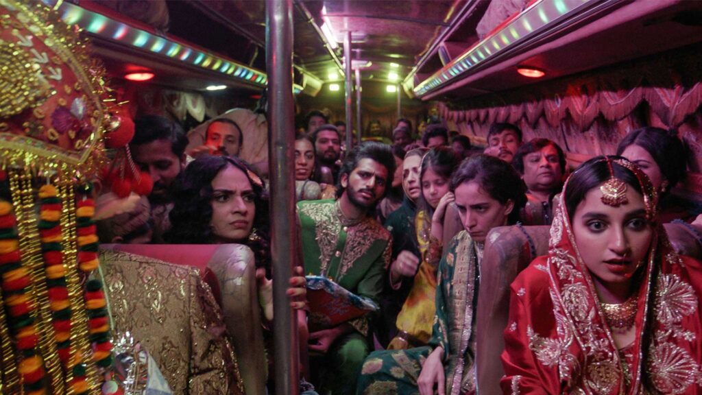 An interior view of a crowd of people on a bus in colorful outfits, a red-clad bride prominent in the front. The lighting is dark and moody, but the image is full of color.