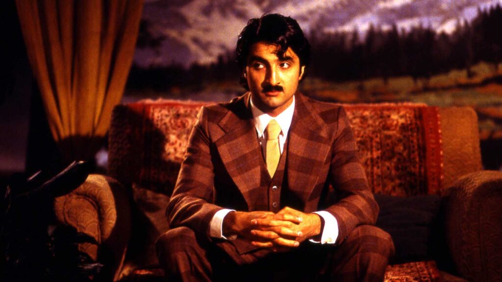 A man in a retro-70's kind of brown paid suit sits on a stylized sofa.